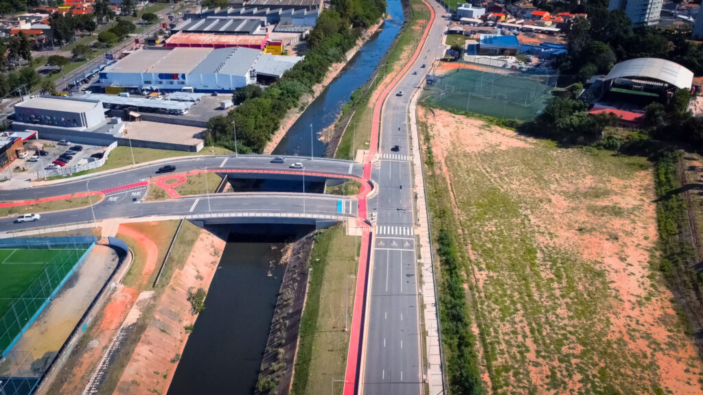 Vista aérea de via urbana com sinalização horizontal completa, incluindo faixas de pedestres, ciclovia, marcações de pare e organização do tráfego sobre ponte e avenida.